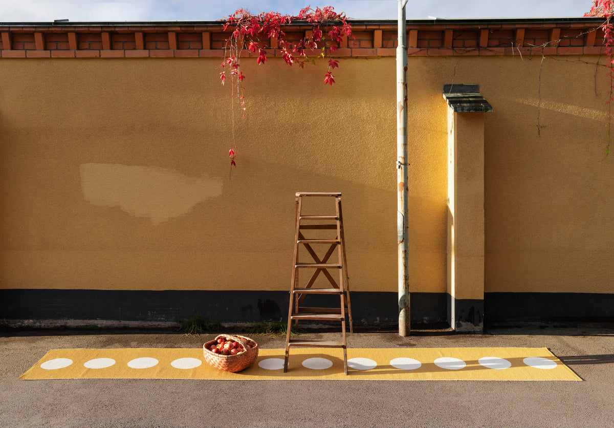 Pappelina VERA woven plastic rug in ochre running across a polished floor in a warm ochre-walled room