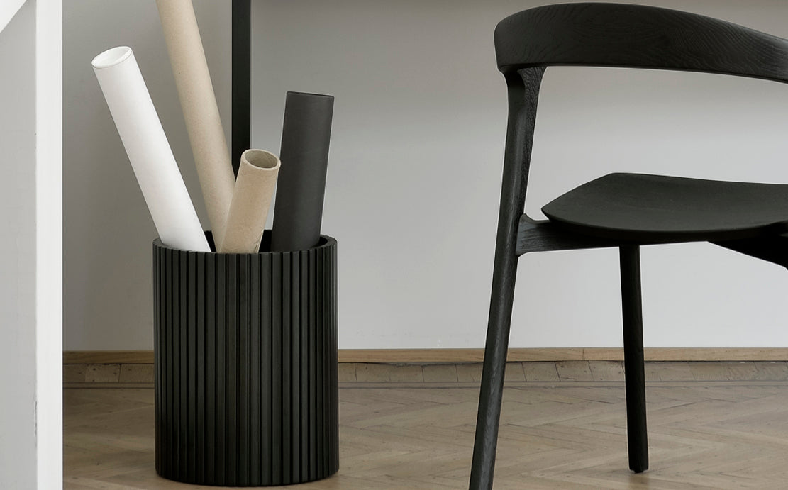 Black Waste Paper Basket by Ethnicraft, showing front view of basket with chair in the live shot.